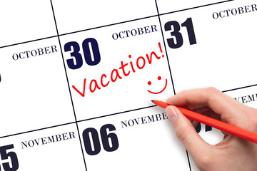 A hand writing a VACATION text and drawing a smiling face on a calendar date 30 October. Vacation...