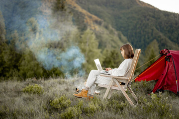 Young woman works on laptop while sitting relaxed on chair by the campfire, traveling with tent in...