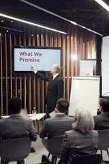 Gray-haired senior business coach in black suit pointing at plasma screen while teaching business...