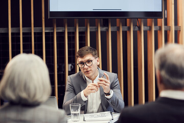 Serious smart young business expert in eyeglasses sitting at table and twisting pen while talking to colleagues at conference - Powered by Adobe