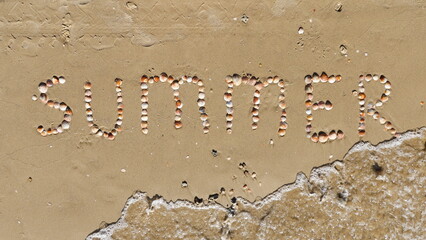 Summer inscription on the sand. The word summer laid out with shells on the sand and a sea wave. Letters made by Shells.  Vacation concept. Seasonal Symbol.