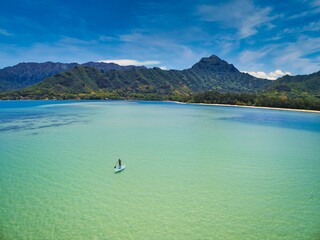 Aerial view of a paddle boarder on open ocean near green mountains on the shore of Oahu, Hawaii