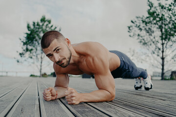 Motivational bearded man stands in plank pose, trains muscles and has strong body. Sporty adult guy performs push up exercise outdoor. Bodybuilder has abdominal workout. Active lifestyle concept