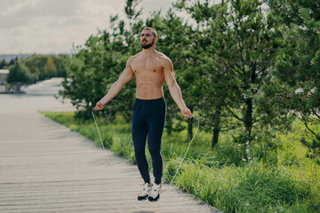 Sporty man with thick beard does exercises with jumping rope, has muscular torso, stays in good physical shape, poses outdoor. Fitness and healthy concept. Male runner warms up with sport equipment