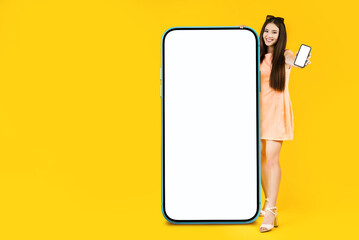 Pretty young asian woman standing near the big smartphone with mockup white screen for your app, text or advertisement design. Blank cellphone in her hand. Studio shoot over the yellow background.