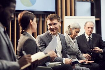 Obraz na płótnie Canvas Smiling handsome young businessman in suit sitting among colleagues at meeting and talking to conference participant cheerfully