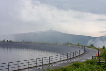 Upper water reservoir of the pumped storage hydro power plant Dlouhe Strane in Jeseniky Mountains, Czech Republic. Top of the Praded mountain behind the lake. Summer foggy  rainy morning, sunrise.