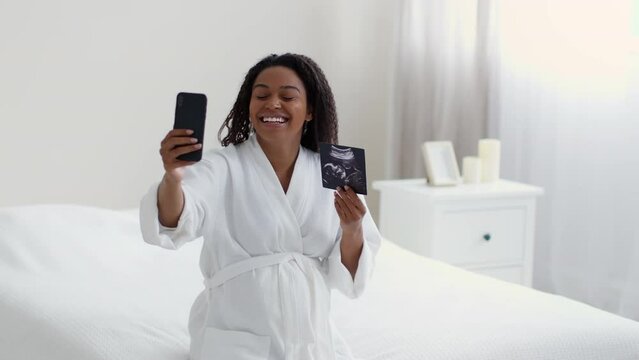 Young happy pregnant lady video chatting with friends via cellphone, showing sonogram and talking to phone, slow motion