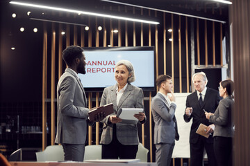 Smiling Asian businesswoman with clipboard standing in modern meeting room and analyzing annual report with black manager at business meeting