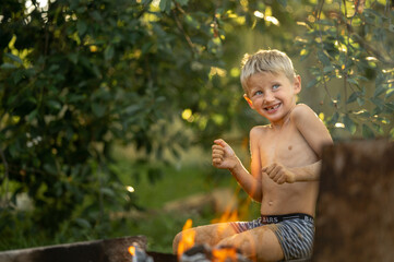 The boy is sitting near the fire, lighting twigs. The child spends the summer holidays in the village.