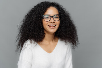Portrait of smiling African American woman wears transparent eyewear and white jumper, being in high spirit, expresses good emotions, isolated over grey background. People and ethnicity concept