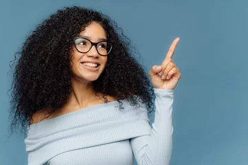 Tuinposter Pleased smiling dark skinned female with Afro hairstyle points index finger upwards, demonstrates something on blank space, has glad expression, wears blue sweater, stands indoor. People and promotion © VK Studio