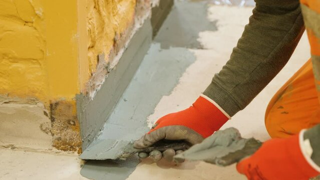 The hands of a worker in red gloves use a spatula to putty the protrusion of the yellow wall with the floor.