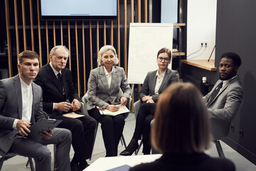 Group of serious multi-ethnic business people of different ages sitting in semi-circle against...