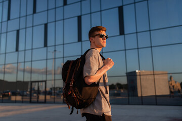 Portrait of handsome tourist hipster man in sunglasses with bag handy, standing guy near the building in downtown. Calm relaxing moment, summer vacation concept - 516445687