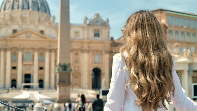 View of the blonde woman from behind in Vatican City. Saint Peter Square with multiple tourists, Saint Peter Basilica and the Vatican Obelisk on the background. Slow motion