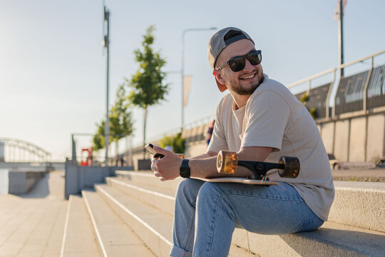 Man sit with skateboard in city landscape.beard guy with skateboard sitting on the street smiling happy. - Powered by Adobe