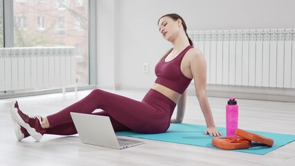 a woman in a burgundy tracksuit with laptop on a mat in a gym. online fitness