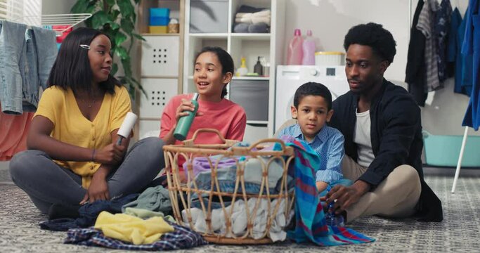 A family sits on the laundry room floor sorting clothes before putting them in the washing machine, playing together during household chores, singing to random objects.