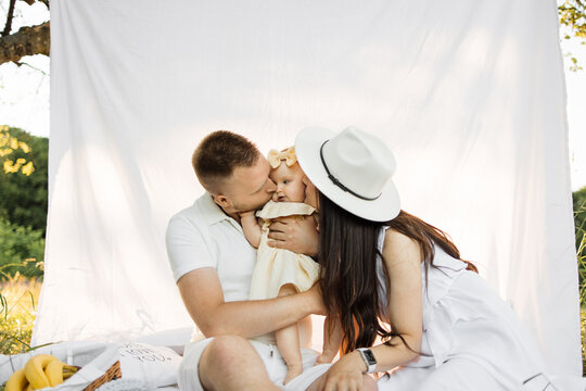 Happy caring mother and father embracing and kissing their little daughter in cheeks. Caucasian family spending summer time on fresh air with fun and relaxation. Copy space.