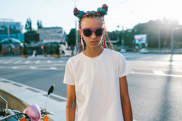 Portrait of a cute teenage girl with African braids in a white t-shirt and sunglasses stands on a...