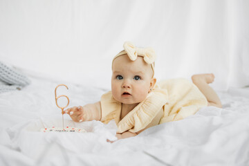 Happy baby with yellow bow on head lying on bed with white background. Smiling infant kid girl in dress lying with cake, child six months old.