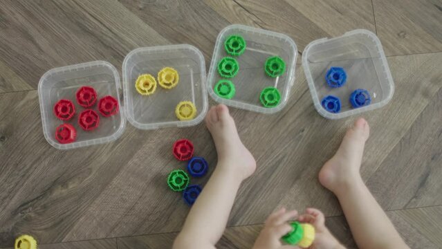 DIY baby educational game learning colors, sorting colorful toys in different boxes. toddler child kid sitting on wooden floor indoors, putting multicolored caps in containers. early education concept