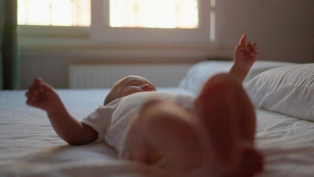 The baby in white lies on the bed and jerks its legs