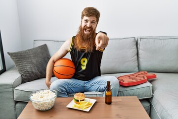 Caucasian man with long beard holding basketball ball cheering tv game pointing to you and the camera with fingers, smiling positive and cheerful