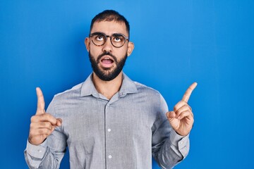 Middle east man with beard standing over blue background amazed and surprised looking up and pointing with fingers and raised arms.