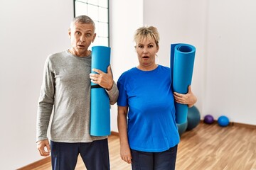 Middle age caucasian couple holding yoga mat at pilates room scared and amazed with open mouth for...