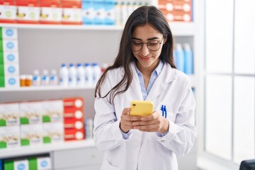 Young hispanic girl pharmacist using smartphone working at pharmacy