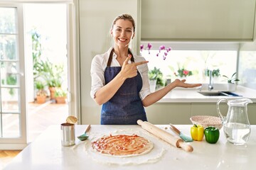 Obraz na płótnie Canvas Beautiful blonde woman wearing apron cooking pizza amazed and smiling to the camera while presenting with hand and pointing with finger.