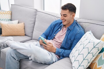 Young hispanic man using smartphone sitting on sofa at home