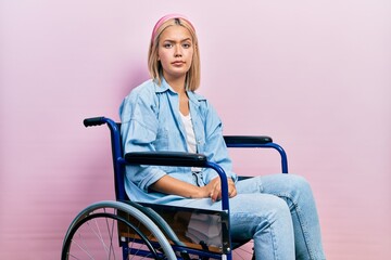 Beautiful blonde woman sitting on wheelchair skeptic and nervous, frowning upset because of...