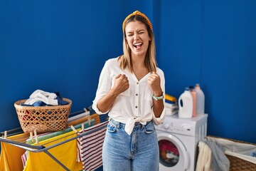 Young blonde woman at laundry room very happy and excited doing winner gesture with arms raised, smiling and screaming for success. celebration concept.