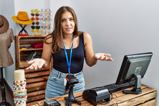 Young brunette woman holding banner with open text at retail shop clueless and confused with open arms, no idea concept.