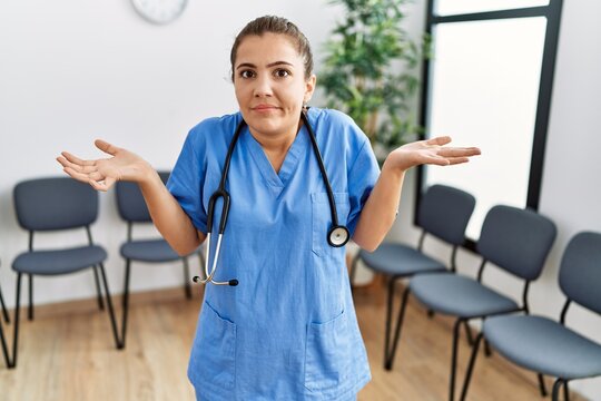 Young brunette doctor woman at waiting room clueless and confused expression with arms and hands raised. doubt concept.