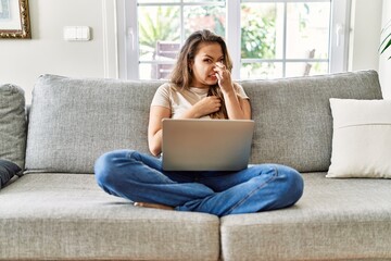 Beautiful young brunette woman sitting on the sofa using computer laptop at home smelling something...