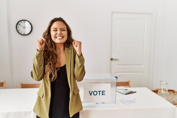 Beautiful hispanic woman standing at political campaign room excited for success with arms raised and eyes closed celebrating victory smiling. winner concept.