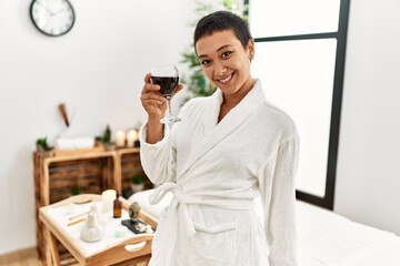 Young hispanic woman wearing bathrobe drinking wine at beauty center