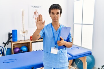 Young hispanic woman with short hair working at pain recovery clinic doing stop sing with palm of...
