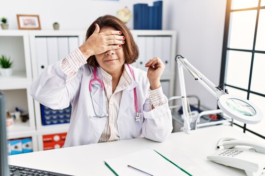 Middle age hispanic woman wearing doctor uniform and stethoscope at the clinic smiling and laughing with hand on face covering eyes for surprise. blind concept.