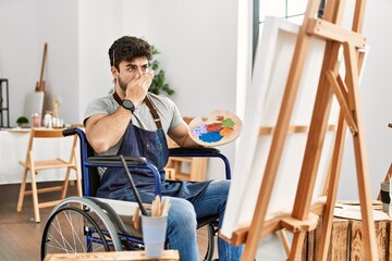 Young hispanic man sitting on wheelchair painting at art studio shocked covering mouth with hands for mistake. secret concept.