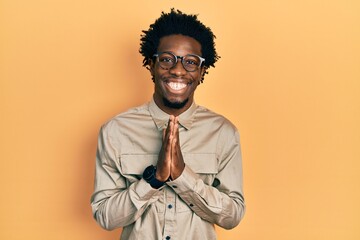 Fototapeta premium Young african american man wearing casual clothes and glasses praying with hands together asking for forgiveness smiling confident.