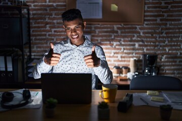 Young hispanic man working at the office at night approving doing positive gesture with hand, thumbs up smiling and happy for success. winner gesture.