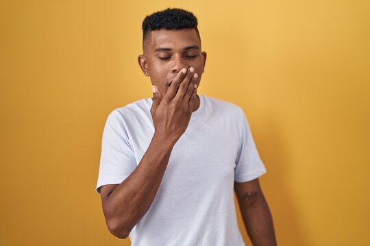 Young hispanic man standing over yellow background bored yawning tired covering mouth with hand. restless and sleepiness.