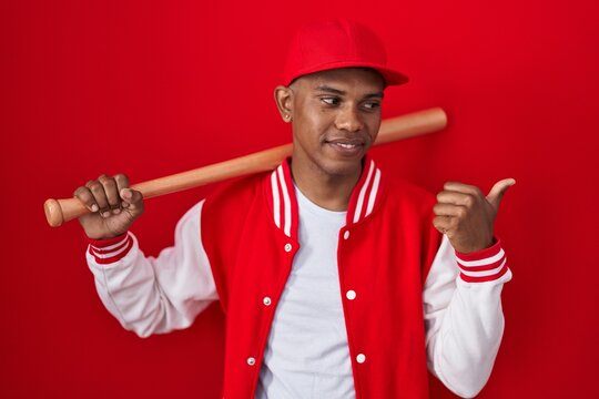 Young hispanic man playing baseball holding bat smiling with happy face looking and pointing to the side with thumb up.