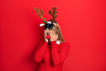 Beautiful hispanic woman wearing deer christmas hat and red nose with sad expression covering face...
