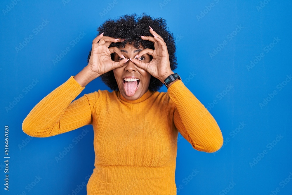 Poster Black woman with curly hair standing over blue background doing ok gesture like binoculars sticking tongue out, eyes looking through fingers. crazy expression.